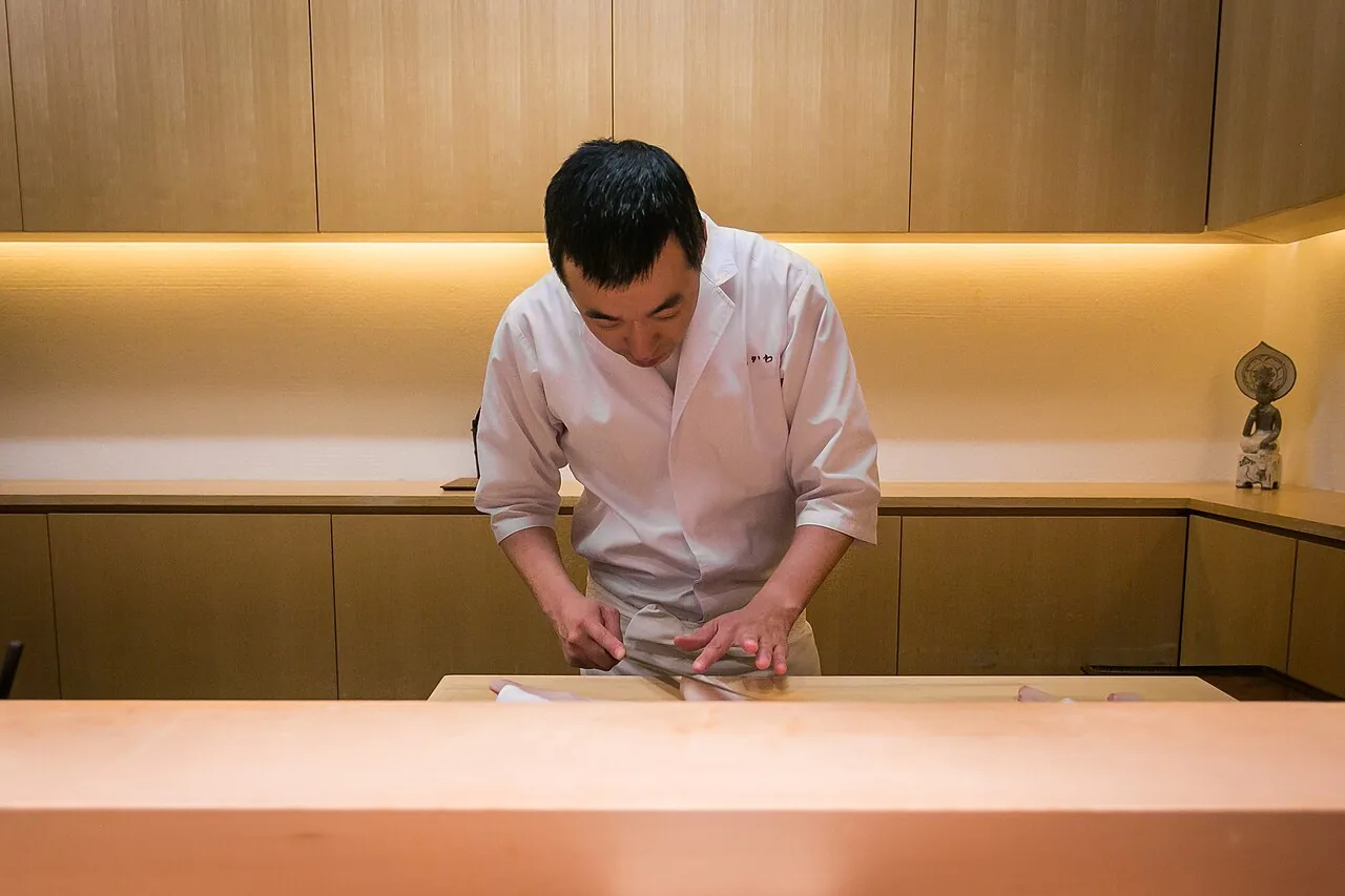 Sushi chef in a white jacket slicing fish at a wooden counter
