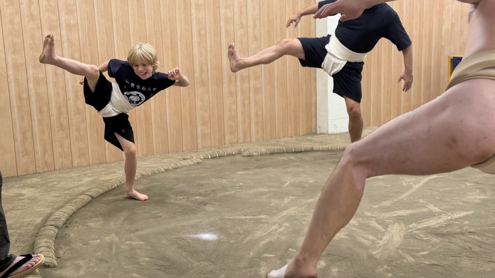 A smiling child raising a leg in the shiko stance beside a wrestler on the dohyo at the Sunamachi Ginza Sumo Stable