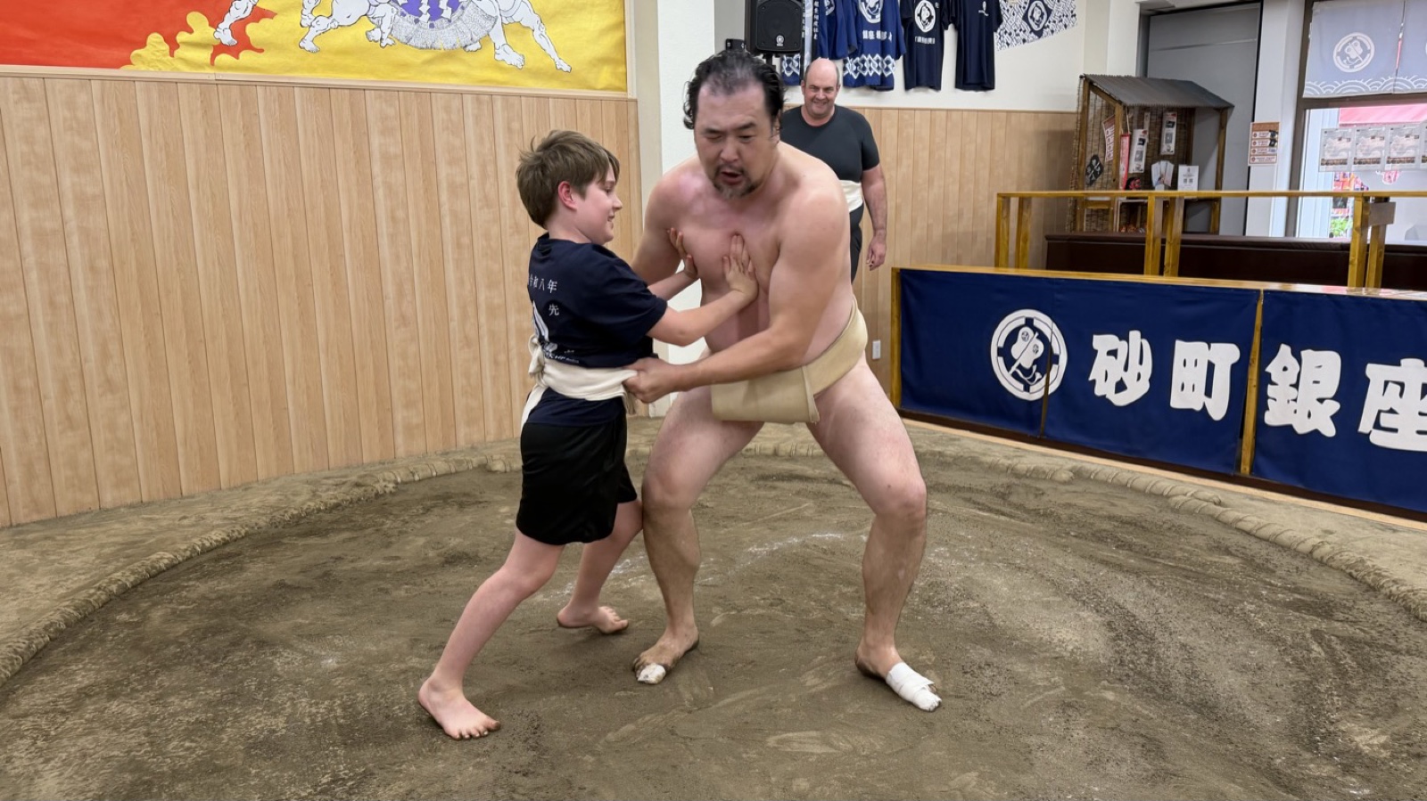 A young guest gripping a wrestler’s mawashi on the dohyo at the Sunamachi Ginza Sumo Stable, with the stable banner behind