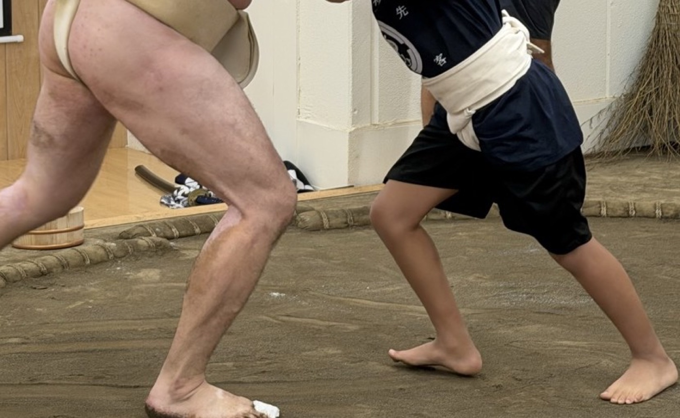 A guest at the Sunamachi Ginza Sumo Stable wearing a white practice mawashi tied over a lent T-shirt and shorts, pushing against a wrestler in a real mawashi on the clay
