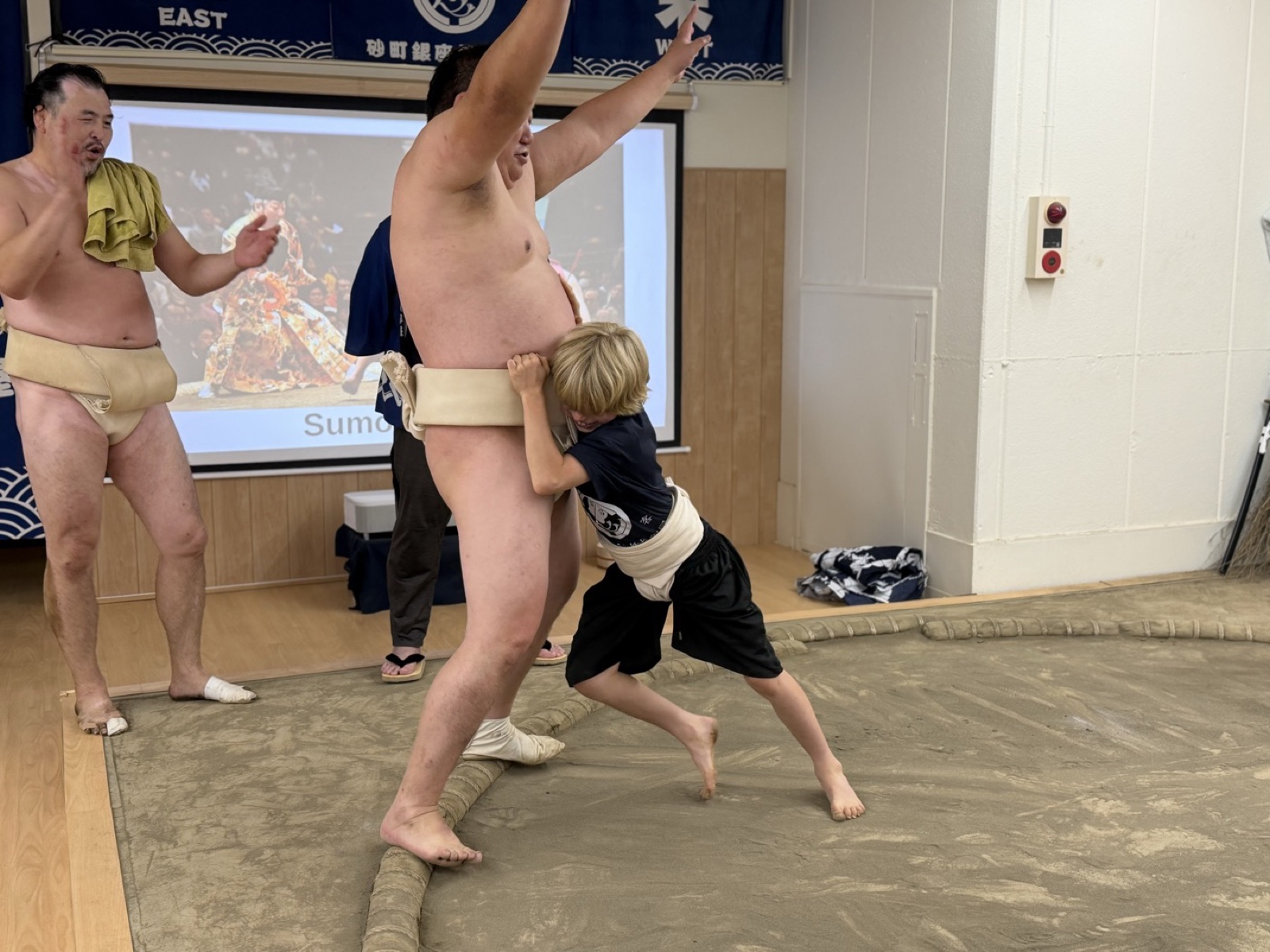 A child driving a wrestler back over the straw tawara bales on the dohyo at the Sunamachi Ginza Sumo Stable